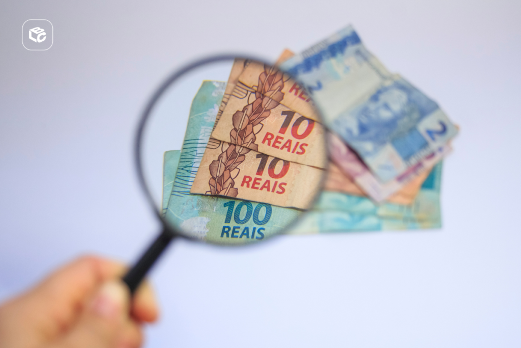 Lupa posicionada sobre cédulas de real brasileiro, ampliando detalhes de notas de R$ 10 e R$ 100. Ao fundo, aparecem outras notas parcialmente sobrepostas, incluindo uma cédula de R$ 2. A imagem destaca a análise ou verificação de valores financeiros, com foco no dinheiro ampliado pela lente. O fundo é claro e desfocado, e há um pequeno logotipo branco no canto superior esquerdo.