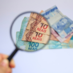 Lupa posicionada sobre cédulas de real brasileiro, ampliando detalhes de notas de R$ 10 e R$ 100. Ao fundo, aparecem outras notas parcialmente sobrepostas, incluindo uma cédula de R$ 2. A imagem destaca a análise ou verificação de valores financeiros, com foco no dinheiro ampliado pela lente. O fundo é claro e desfocado, e há um pequeno logotipo branco no canto superior esquerdo.
